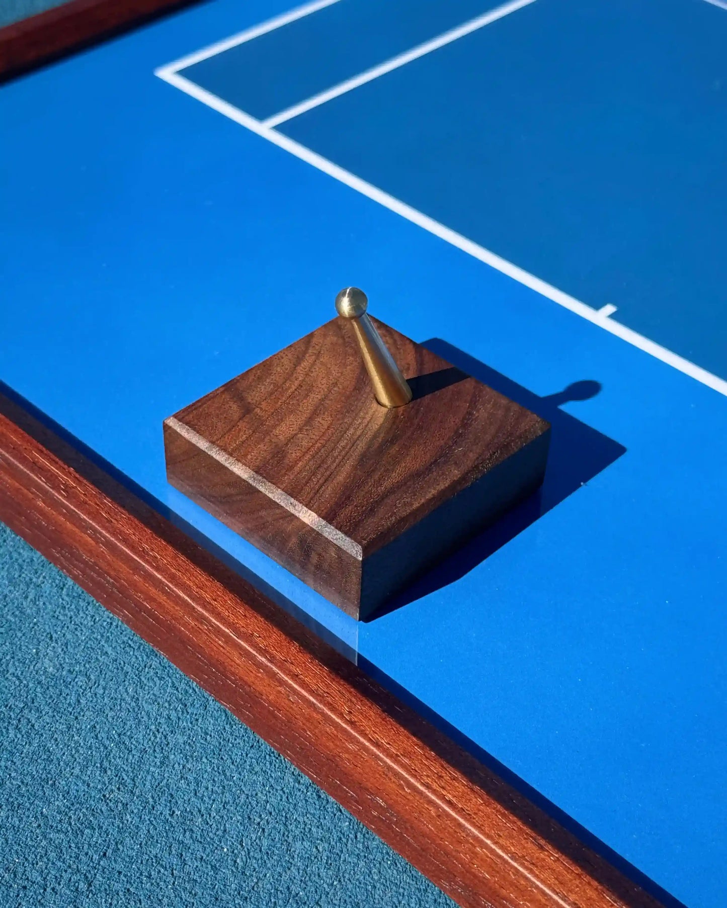 Close-up of Tennis Frame hardware showing walnut hook with brass peg and wood frame edge against blue court background