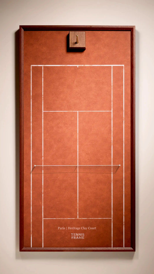A framed tennis court print featuring a terracotta-colored court design with white boundary lines and a dark brown wooden frame, mounted on a light beige wall.
